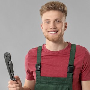 Ein junger Mann mit Bart lächelt freundlich, während er einen Rohrzange in der Hand hält und grüne Latzhose trägt.